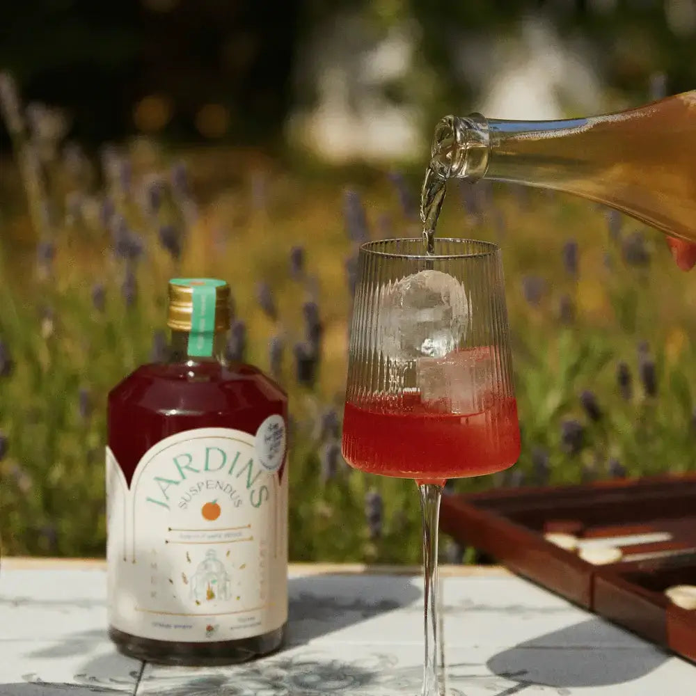 Glas mit rotem alkoholfreiem Bitterorange-Rosmarin-Drink, Eiswürfel, Flasche neben Glas auf weißem Tisch, Gartenhintergrund.